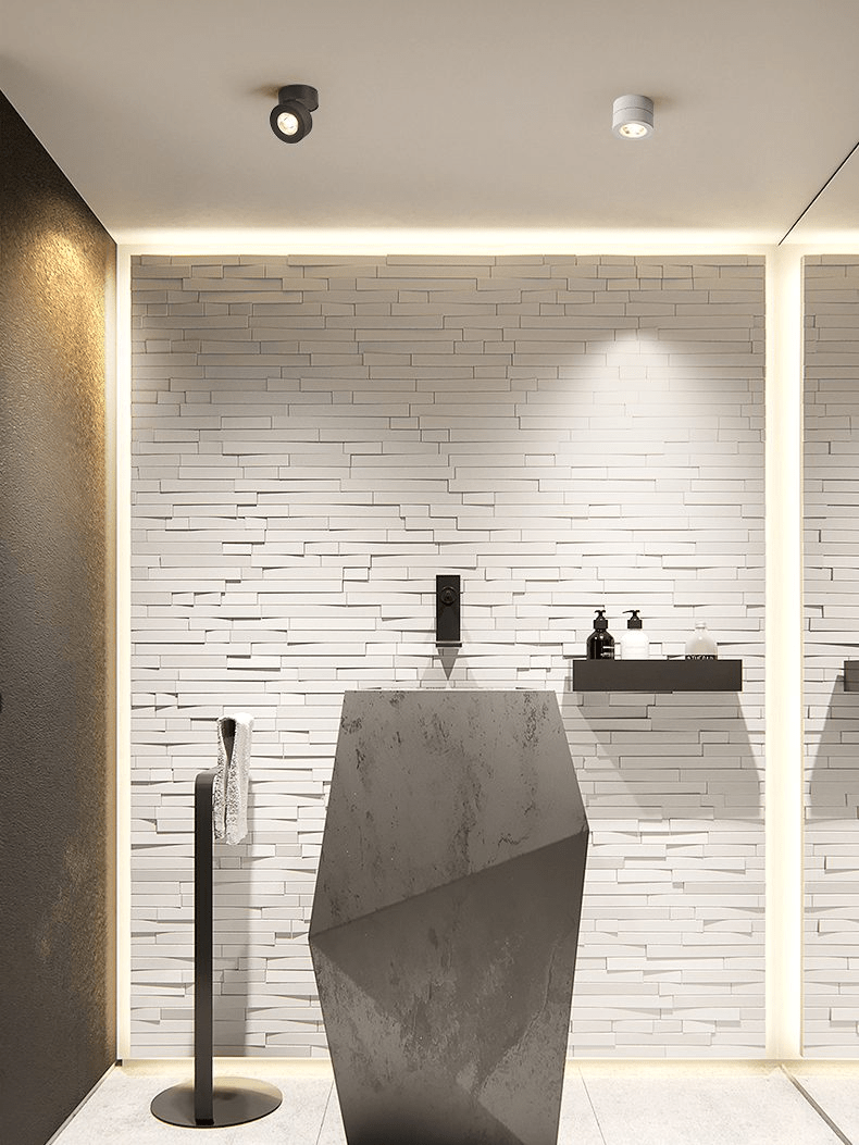 A black and a white Clérachic spotlight on the ceiling of a washroom, with a warm light strip along a brick-effect wall and a stone basin.