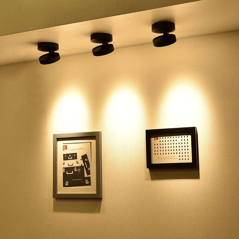 Three black Clérachic spotlights tilted at a cream wall, each casting a warm pool of light beside two framed pictures.