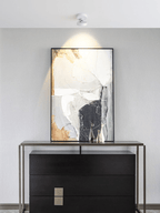 A Clérachic spotlight on the ceiling, lighting a large abstract painting above a black chest of drawers on a pale grey wall.