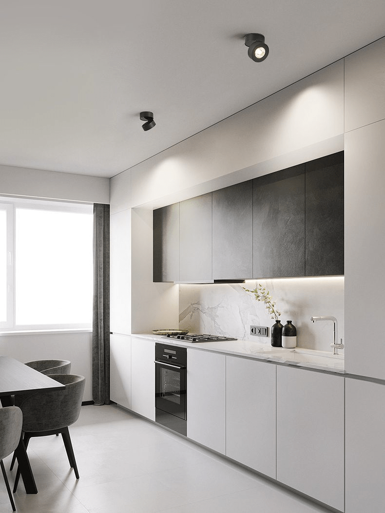 Black Clérachic spotlights on the ceiling of a modern kitchen, one tilted, above grey and white cabinets and a dark dining chair.