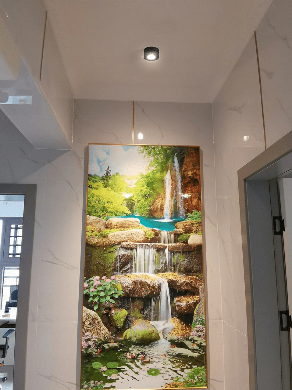A dark Clérachic spotlight on the ceiling of a narrow white hallway, above a framed waterfall landscape picture.