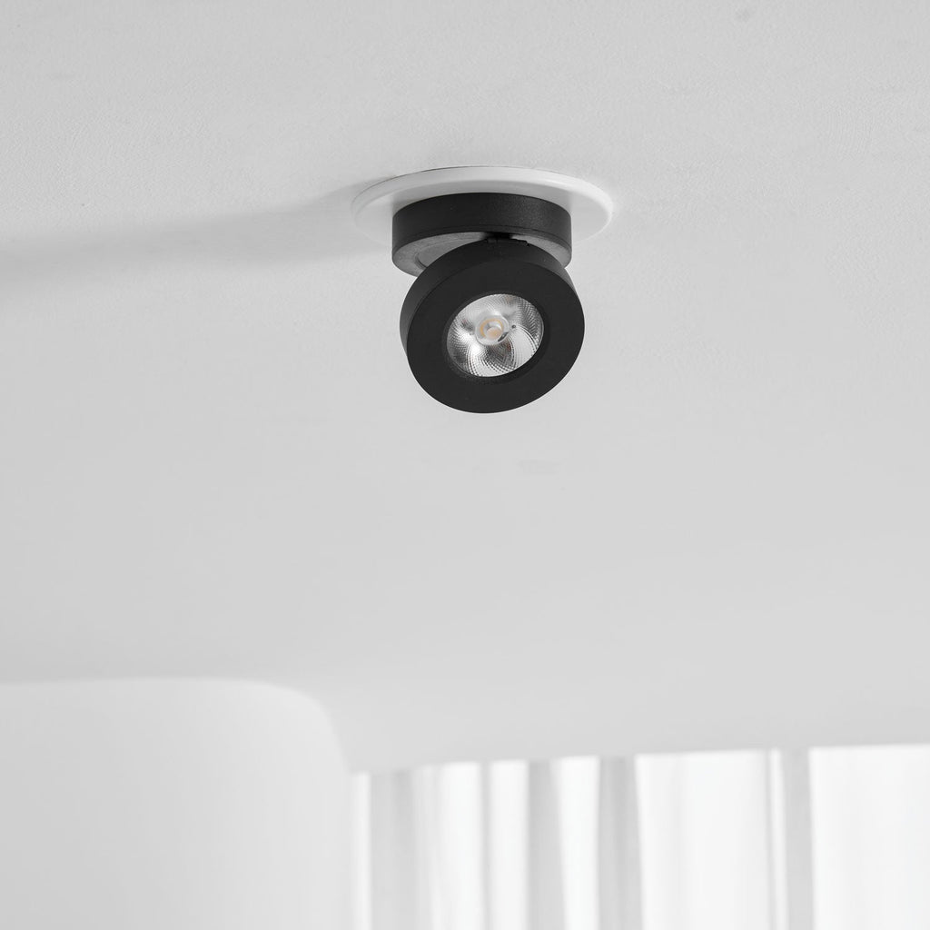 Black Clérachic spotlight set in a white ring on a white ceiling, with its small round head angled to one side and sheer curtains below.