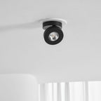 Black Clérachic spotlight set in a white ring on a white ceiling, with its small round head angled to one side and sheer curtains below.