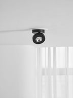 Small, distant view of a black Clérachic spotlight in a white ring on a ceiling, above a window with white curtains.