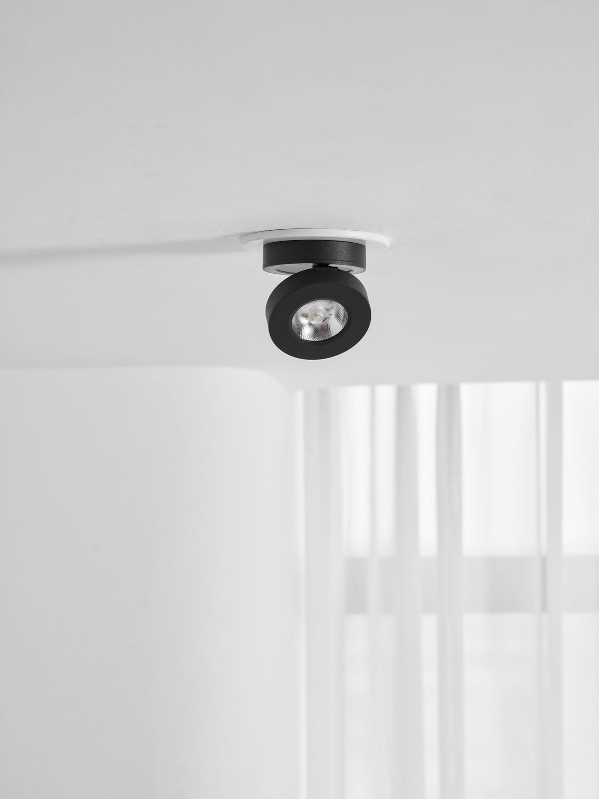 Small, distant view of a black Clérachic spotlight in a white ring on a ceiling, above a window with white curtains.