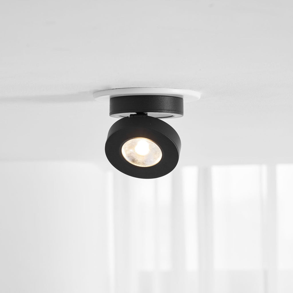 Black Clérachic spotlight in a white ceiling ring, its head hanging low and its LED glowing warm, with a window and curtains behind.