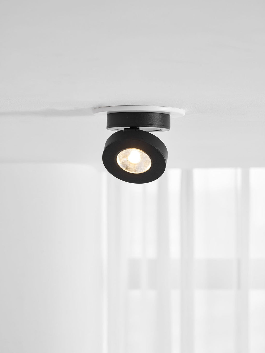 Black Clérachic spotlight with a lit LED, tilted slightly in a white ring on a ceiling, with pale curtains below.