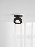 Black Clérachic spotlight with a lit LED, tilted slightly in a white ring on a ceiling, with pale curtains below.