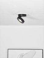 Black Clérachic spotlight angled sharply on a white ceiling, with the corner of a framed line drawing visible below.