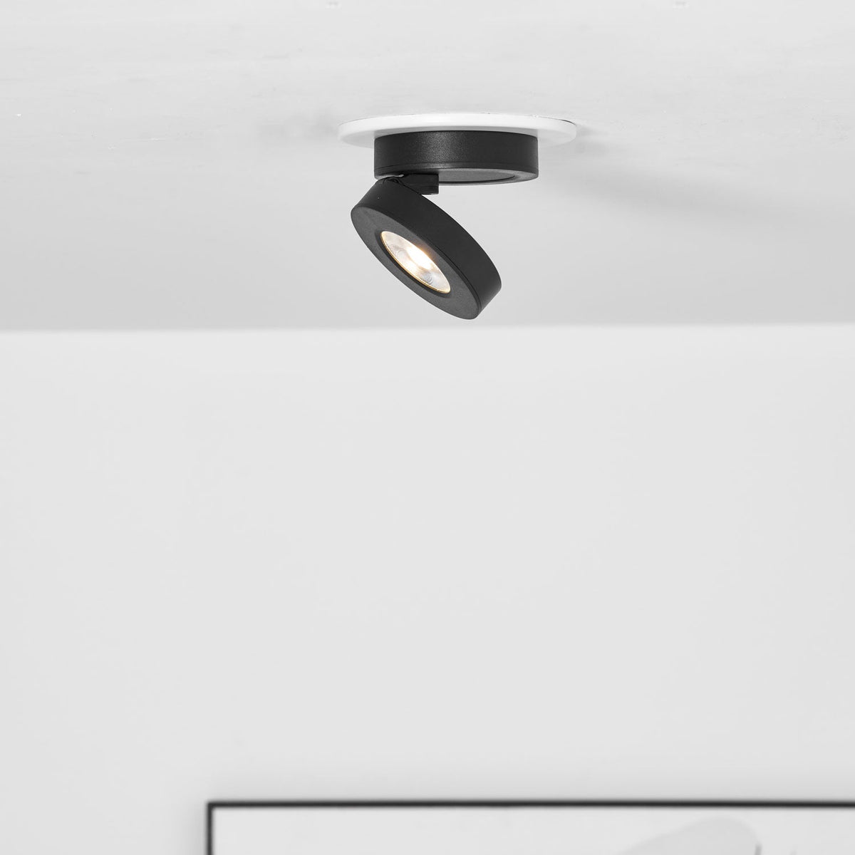 Clérachic black spotlight with its head tipped to one side inside a white ring, above the edge of a framed picture.