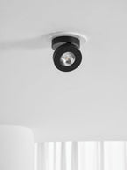 Distant view of a black Clérachic spotlight in a white ceiling ring, above a white wall and the edge of a window curtain.