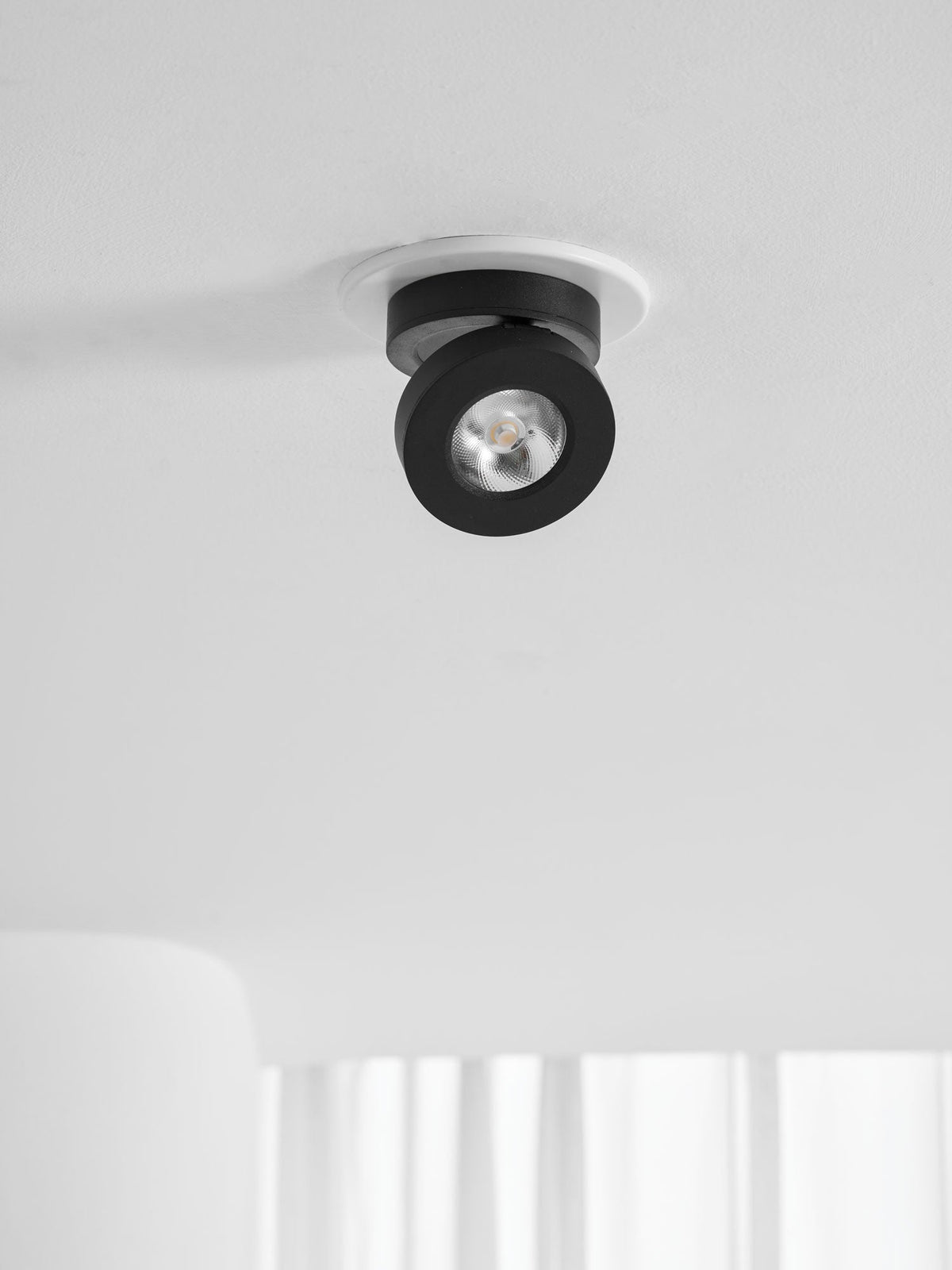Distant view of a black Clérachic spotlight in a white ceiling ring, above a white wall and the edge of a window curtain.