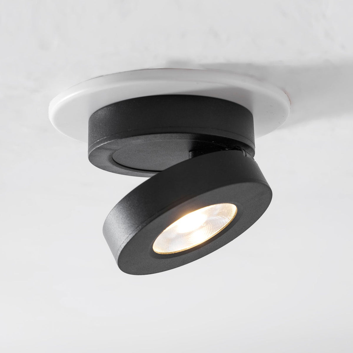 Black Clérachic spotlight with its head tilted well out from the base and its LED lit warm, set into a white ceiling ring.