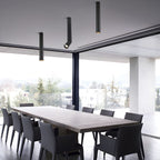 Three black Clérachic spotlights hang at different lengths over a long dining table, in front of floor-to-ceiling glass.