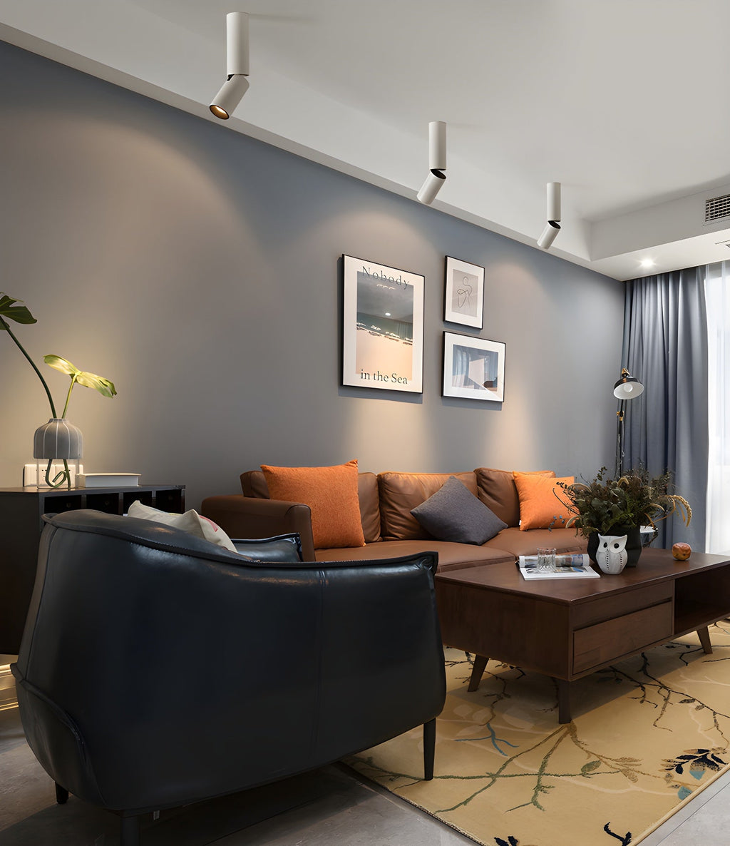 A row of white Clérachic spotlights runs across the ceiling over a leather armchair and a sofa with orange cushions.