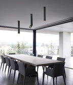 Black Clérachic pole spotlights in three lengths hang above a long table with dark chairs, beside a glass wall.