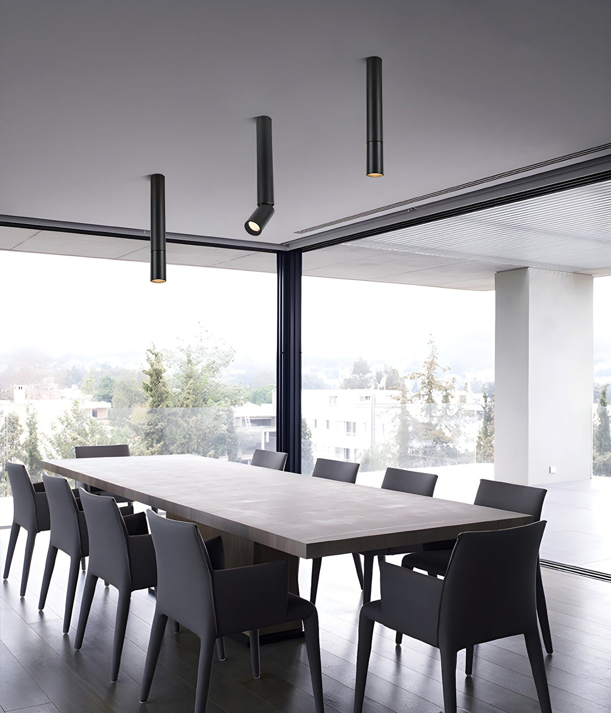 Black Clérachic pole spotlights in three lengths hang above a long table with dark chairs, beside a glass wall.