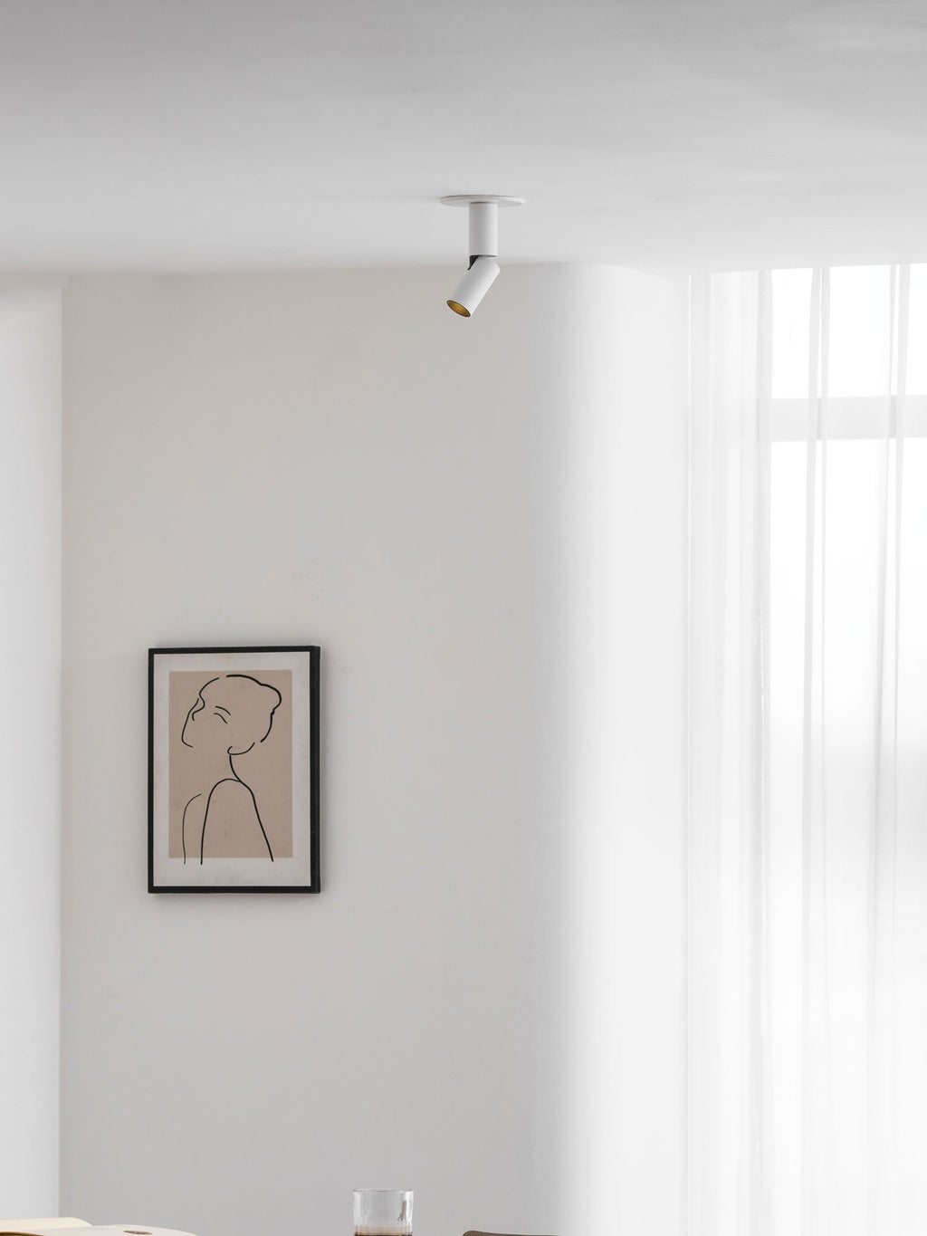 A small white Clérachic spotlight high on a ceiling beside a window with sheer curtains, above a framed line drawing.