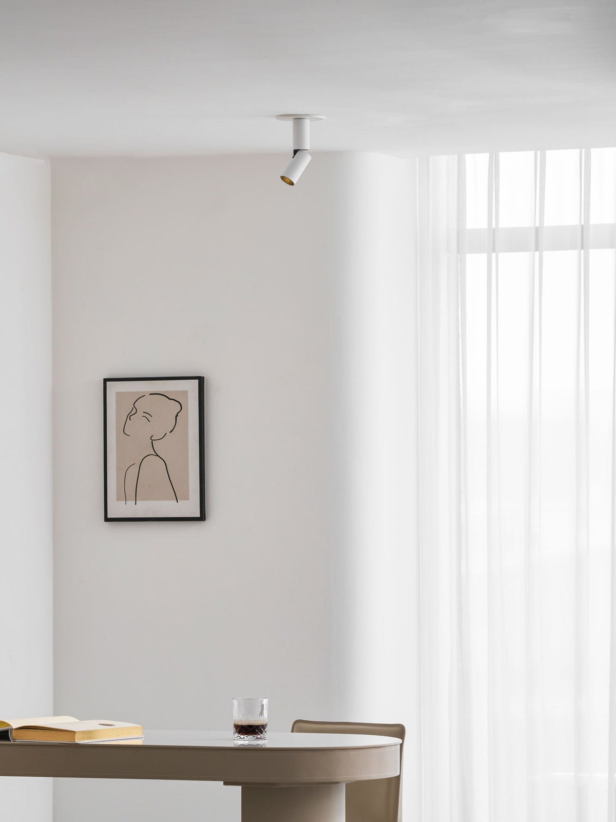 Tiny from a distance, this white Clérachic spotlight sits on a ceiling above a framed print and a table with a book and glass.