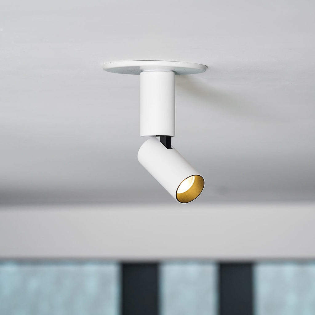 Close view of a white Clérachic spotlight with a black joint and a gold-toned lens ring, with a blurred shelf of bottles behind.