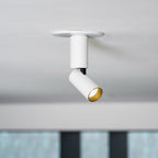 Close view of a white Clérachic spotlight with a black joint and a gold-toned lens ring, with a blurred shelf of bottles behind.