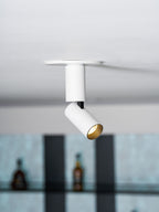 This white Clérachic spotlight tilts its head to one side below a flat ceiling plate, with bottles on shelves out of focus.
