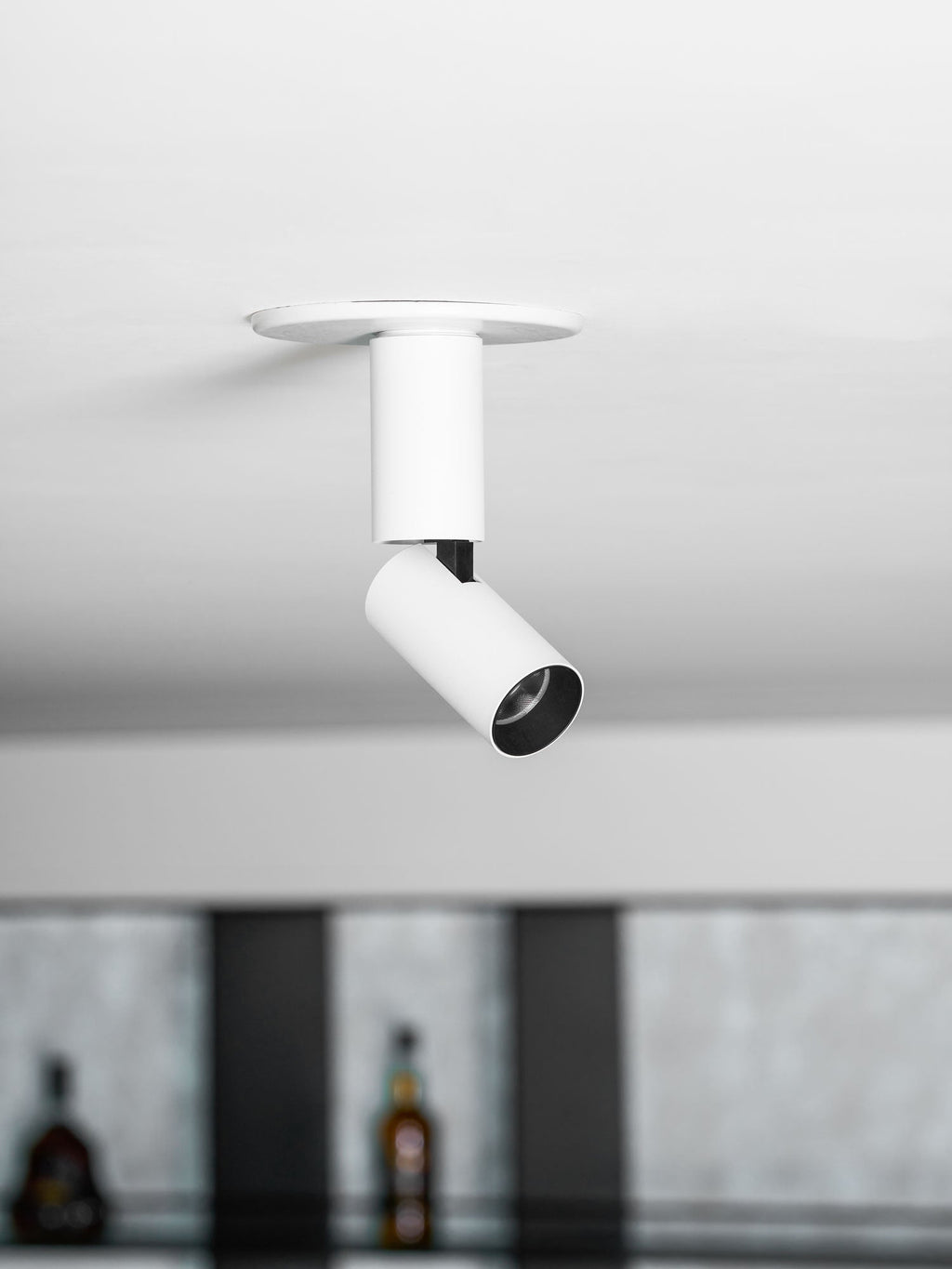 Looking up at a white Clérachic spotlight with a black lens ring and a black swivel joint, with bottles blurred in the background.