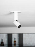 Looking up at a white Clérachic spotlight with a black lens ring and a black swivel joint, with bottles blurred in the background.