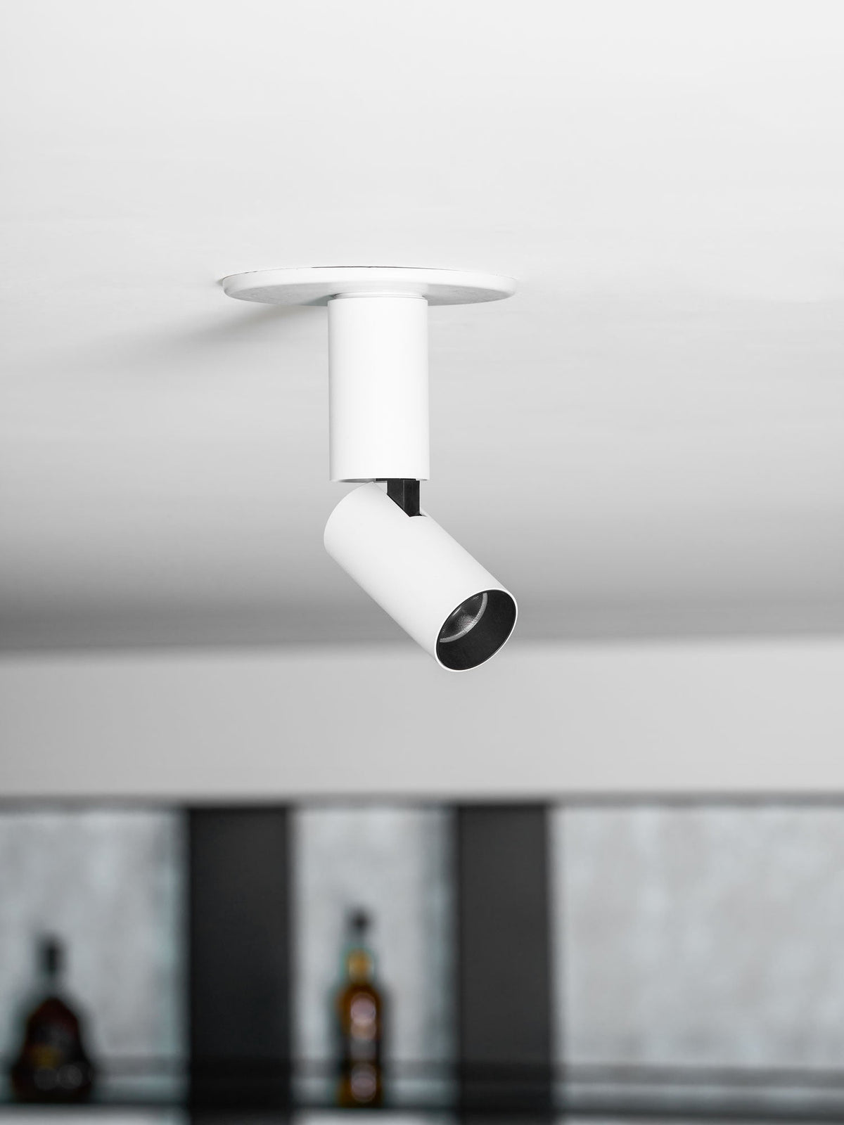 Looking up at a white Clérachic spotlight with a black lens ring and a black swivel joint, with bottles blurred in the background.