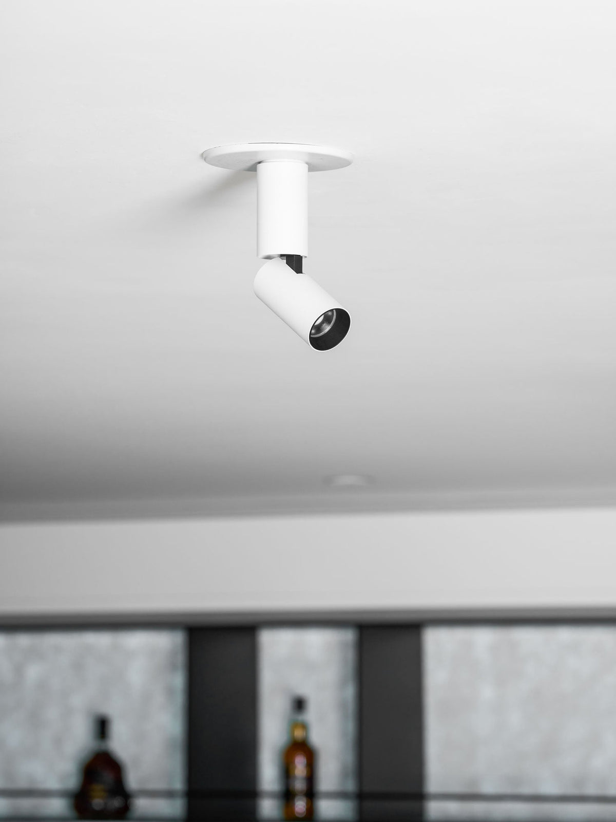 A smaller view of this white Clérachic spotlight, its black lens ring and joint visible, beside blurred shelves of bottles.