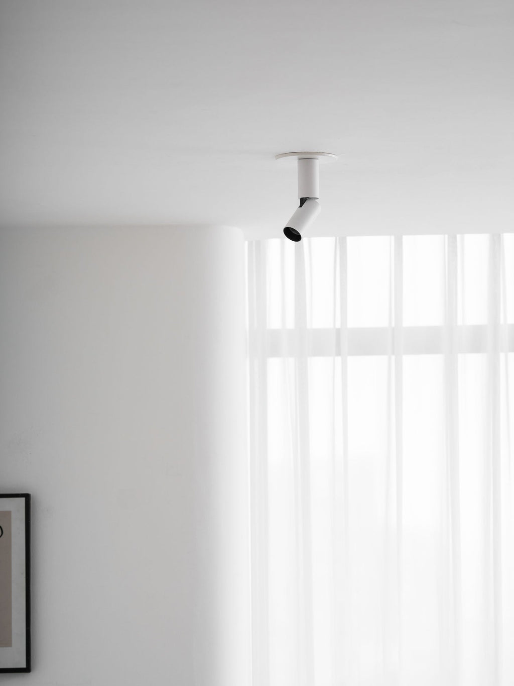 A white Clérachic spotlight high in the corner of a room, its head angled towards the window with sheer curtains.
