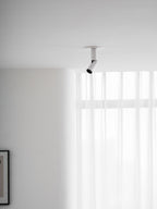 A white Clérachic spotlight high in the corner of a room, its head angled towards the window with sheer curtains.