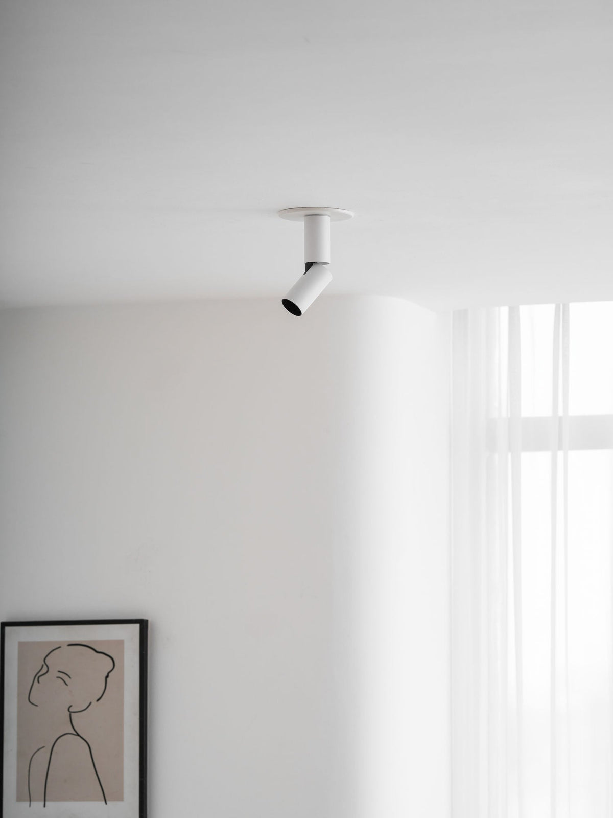 A white Clérachic spotlight on the ceiling, its head pointing down towards the left, above a framed line drawing.