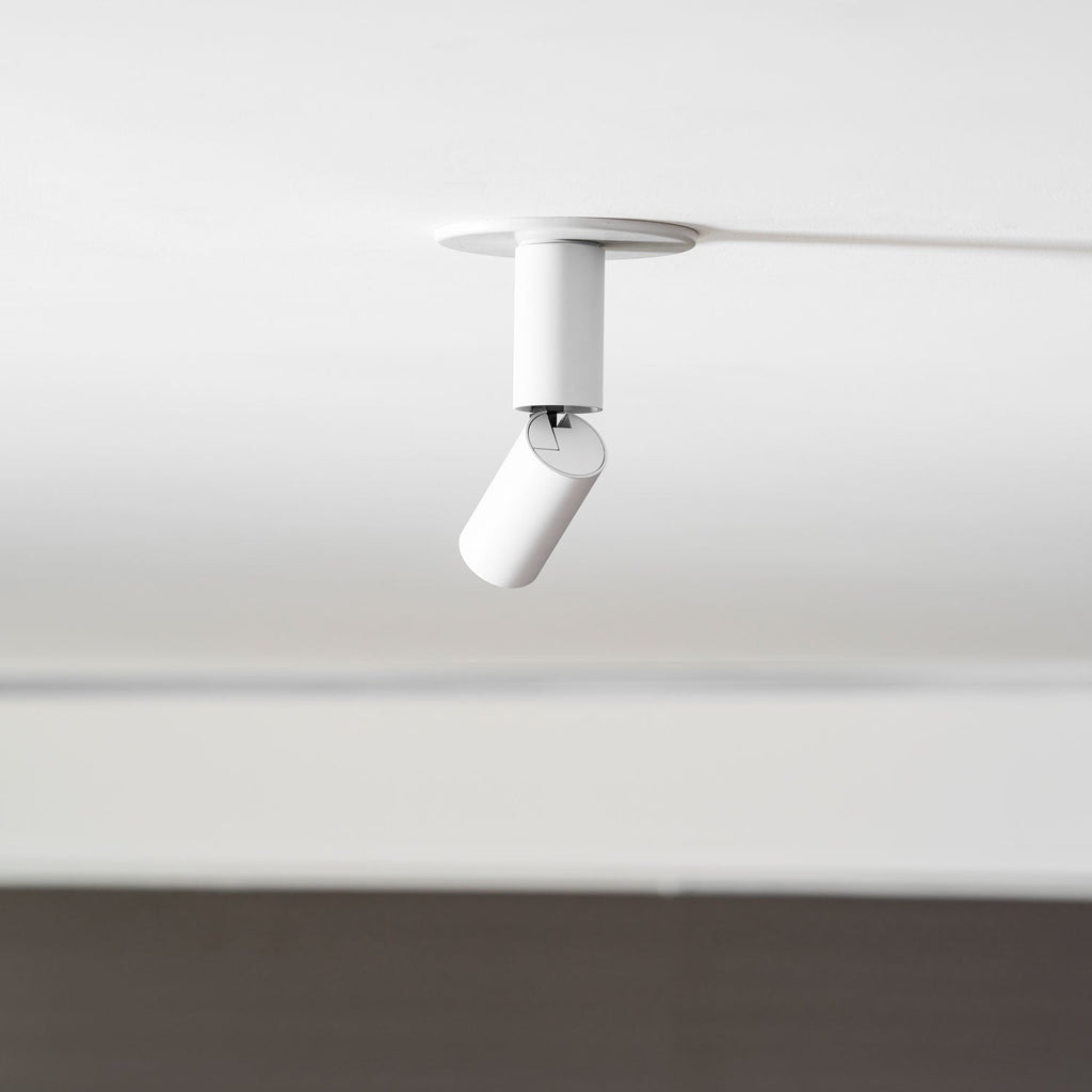 A white Clérachic spotlight with its head swung out at an angle, set against a white ceiling and a dark wall below.