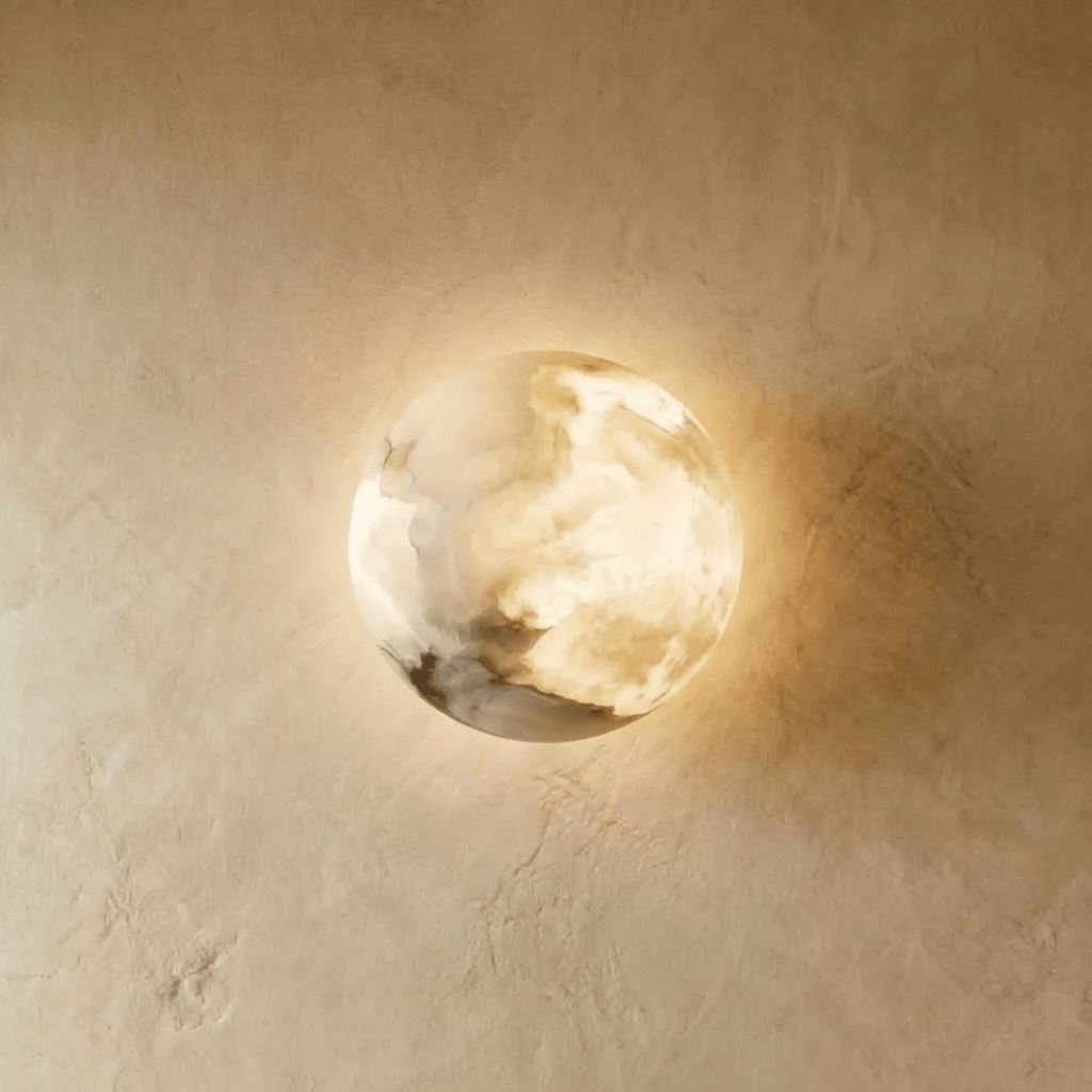 Clérachic alabaster wall lamp, a domed disc of cloudy white veined stone glowing warm on a limewashed plaster wall.