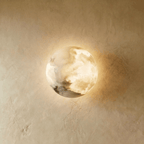 Clérachic alabaster wall lamp, a domed disc of cloudy white veined stone glowing warm on a limewashed plaster wall.