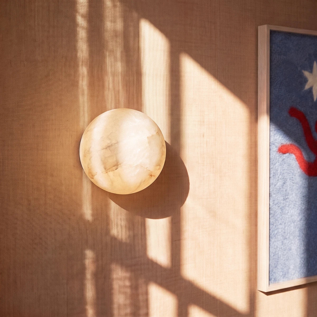 Clérachic alabaster wall lamp with veined cream stone, set in low sun and window shadows on a woven wall covering.