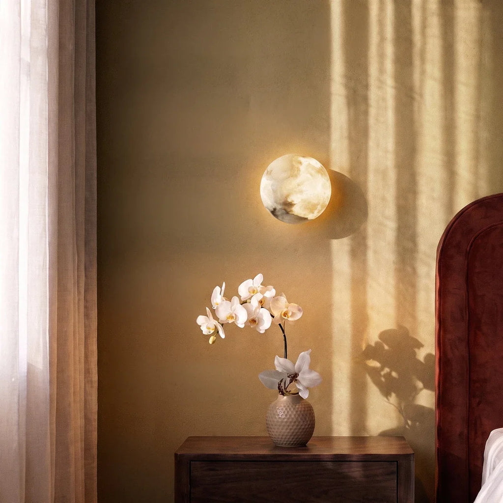Clérachic alabaster wall lamp glowing above a walnut bedside table and a vase of white orchids in warm evening light.