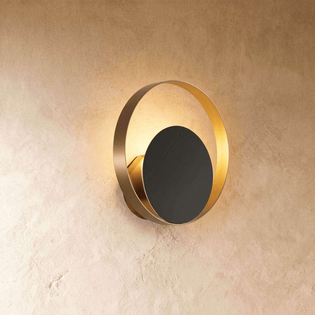 Clérachic round wall lamp with a brushed gold ring and a dark disc in front, warm light glowing round the edge on limewash.