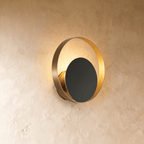 Clérachic round wall lamp with a brushed gold ring and a dark disc in front, warm light glowing round the edge on limewash.