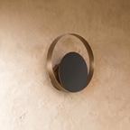Clérachic round wall lamp with an antique-gold ring and a black disc, unlit, mounted on a textured limewash wall in the studio.