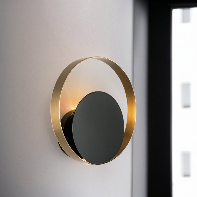 Close up of a Clérachic round wall lamp, a brushed gold ring around a glossy black disc, beside a window in a room.