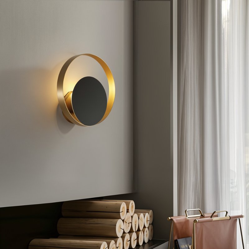 Clérachic round gold ring wall lamp with a black disc glowing warm in a room, above stacked birch logs and sheer curtains.