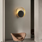 Clérachic round gold ring wall lamp with a black disc lit warmly on a grey wall above a tan velvet armchair in a room.