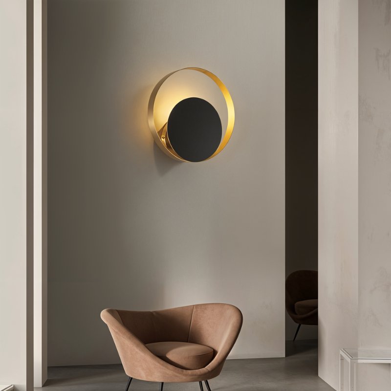 Clérachic round gold ring wall lamp with a black disc lit warmly on a grey wall above a tan velvet armchair in a room.