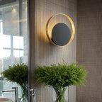 Clérachic round gold ring wall lamp with a black disc on a woven textured wall above a glass vase of green ferns in a room.