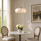 Over a small marble table with two cane-backed chairs, this Clérachic chandelier glows in a cream room with a tall window and framed landscape.