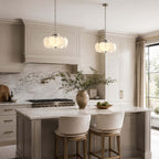 Two Clérachic chandeliers hang in a pale kitchen above a marble-topped island with two upholstered stools and a marble splashback.