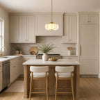 A small Clérachic chandelier with a brass chain hangs over a kitchen island with two cream stools, in front of white cabinets.