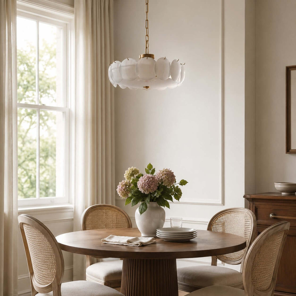 With its white discs lit softly, this Clérachic chandelier hangs from a brass chain over a round table set with pink hydrangeas.
